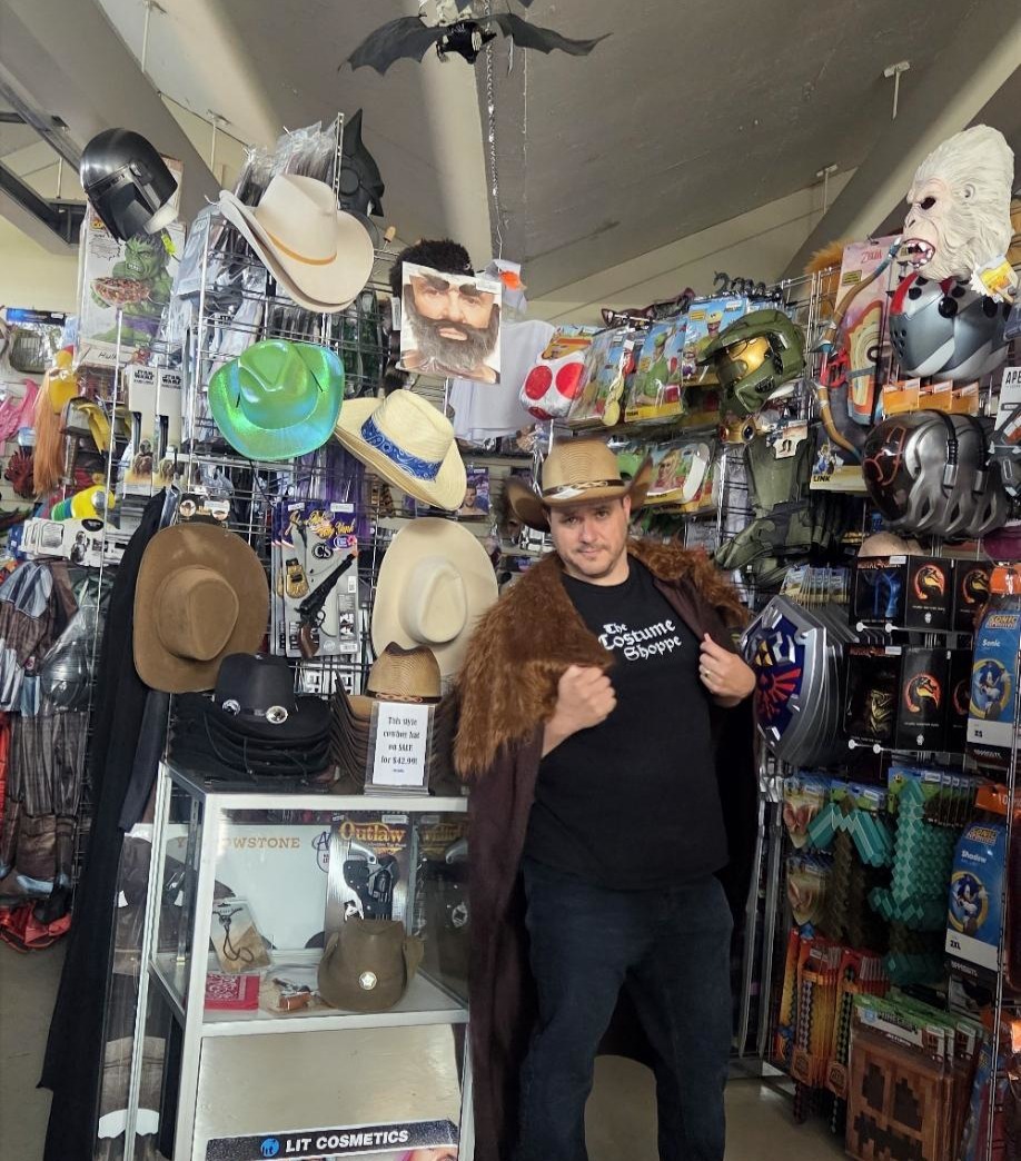 The Costume Shoppe Owner Ry — Cowboy Hat to Cape Calgary Ry The Costume Shoppe Guy, owner of The Costume Shoppe Calgary, wearing a cowboy hat and cape surrounded by western accessories and Halloween costumes at 4307 Blackfoot Trail SE